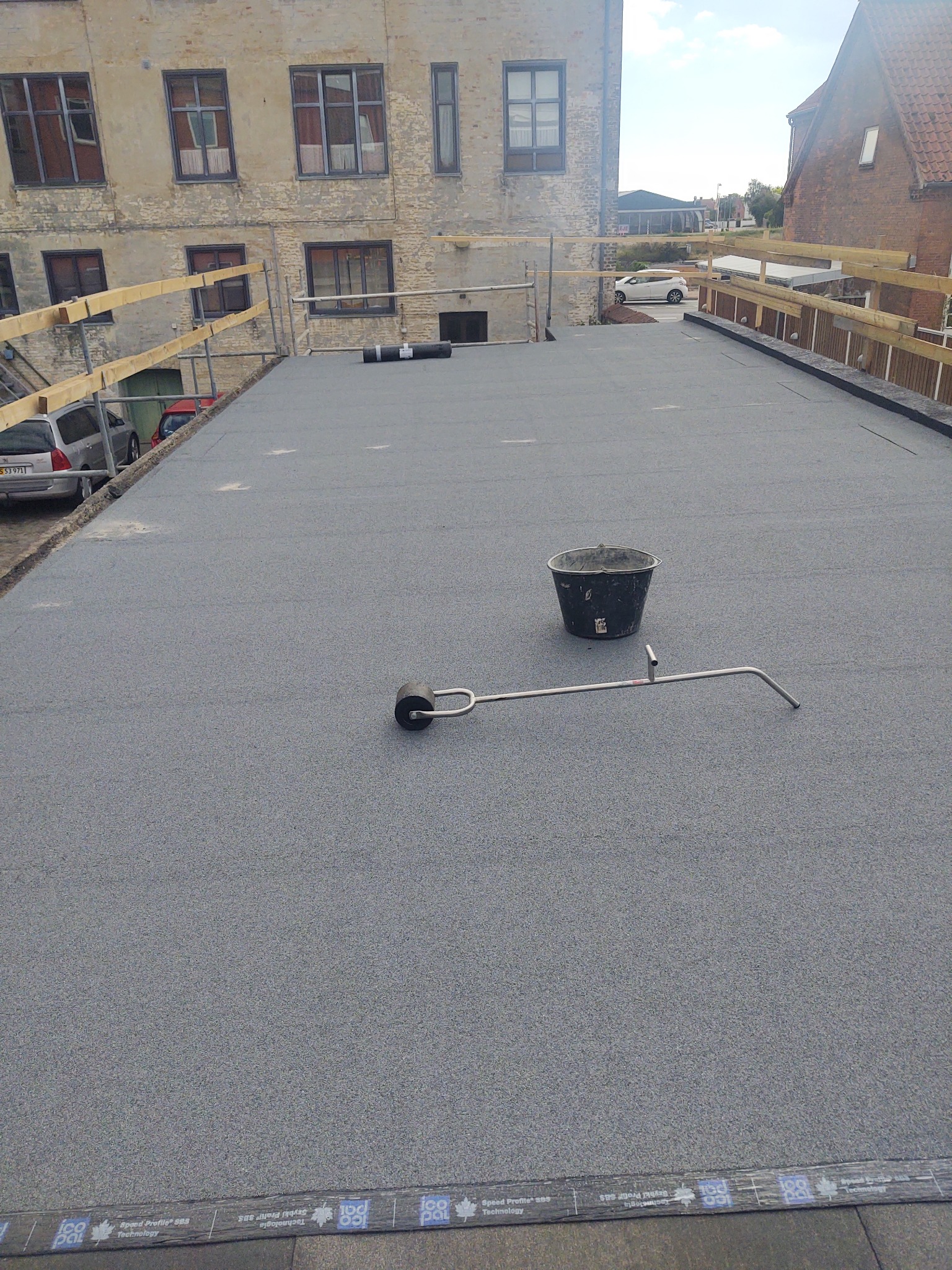 Szary, płaski dach pokryty papą z widocznym wiadrem, wałkiem dociskowym i fragmentem budynku w tle z drewnianą barierką zabezpieczającą.