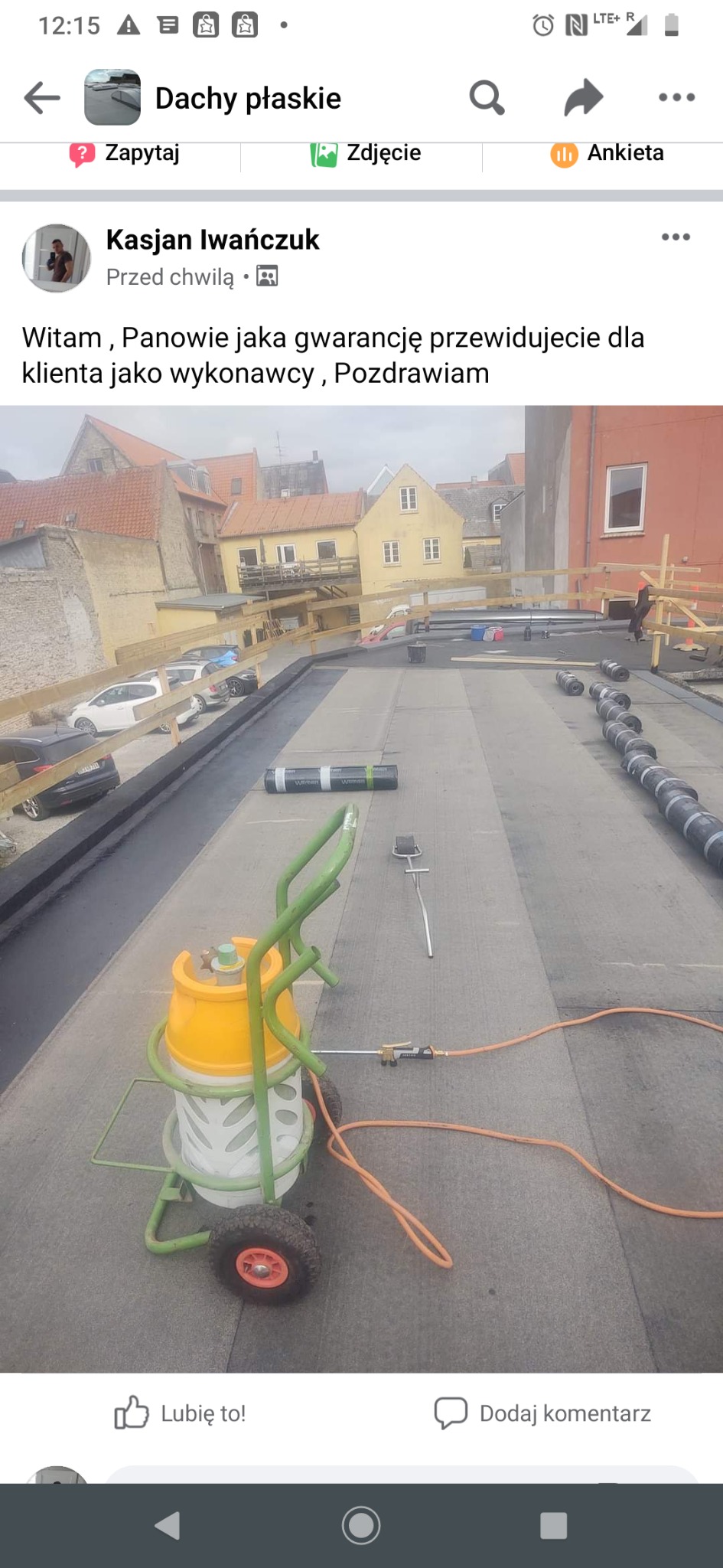 Prace na płaskim dachu: widoczny sprzęt do zgrzewania papy na wózku z butlą gazową, rozłożone rolki papy oraz częściowo pokryta powierzchnia dachu, w tle zabudowa miejska z ceglanymi dachami.