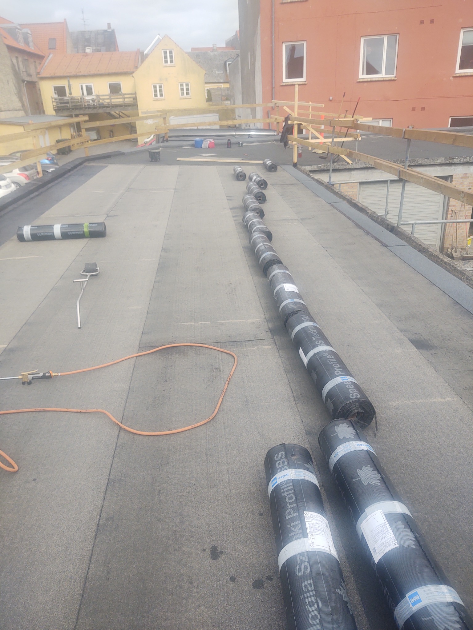 Płaski dach w trakcie pokrywania papą termozgrzewalną, widoczne rolki papy ułożone w rzędzie, palnik dekarski z wężem oraz fragment rusztowania na tle zabudowy miejskiej.