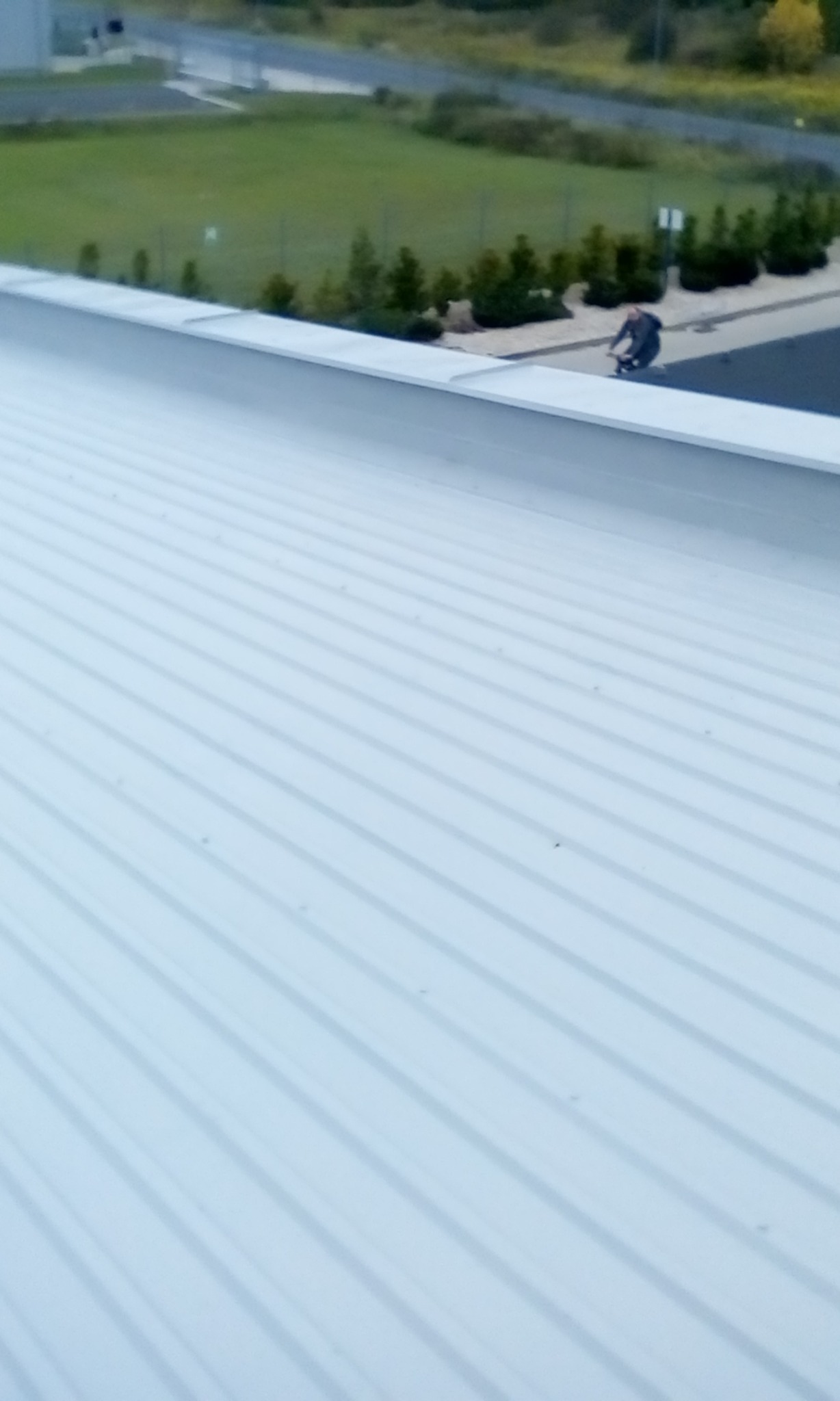 Widok z góry na biały, blaszany dach z widocznym fragmentem chodnika i przechodzącą osobą w tle.