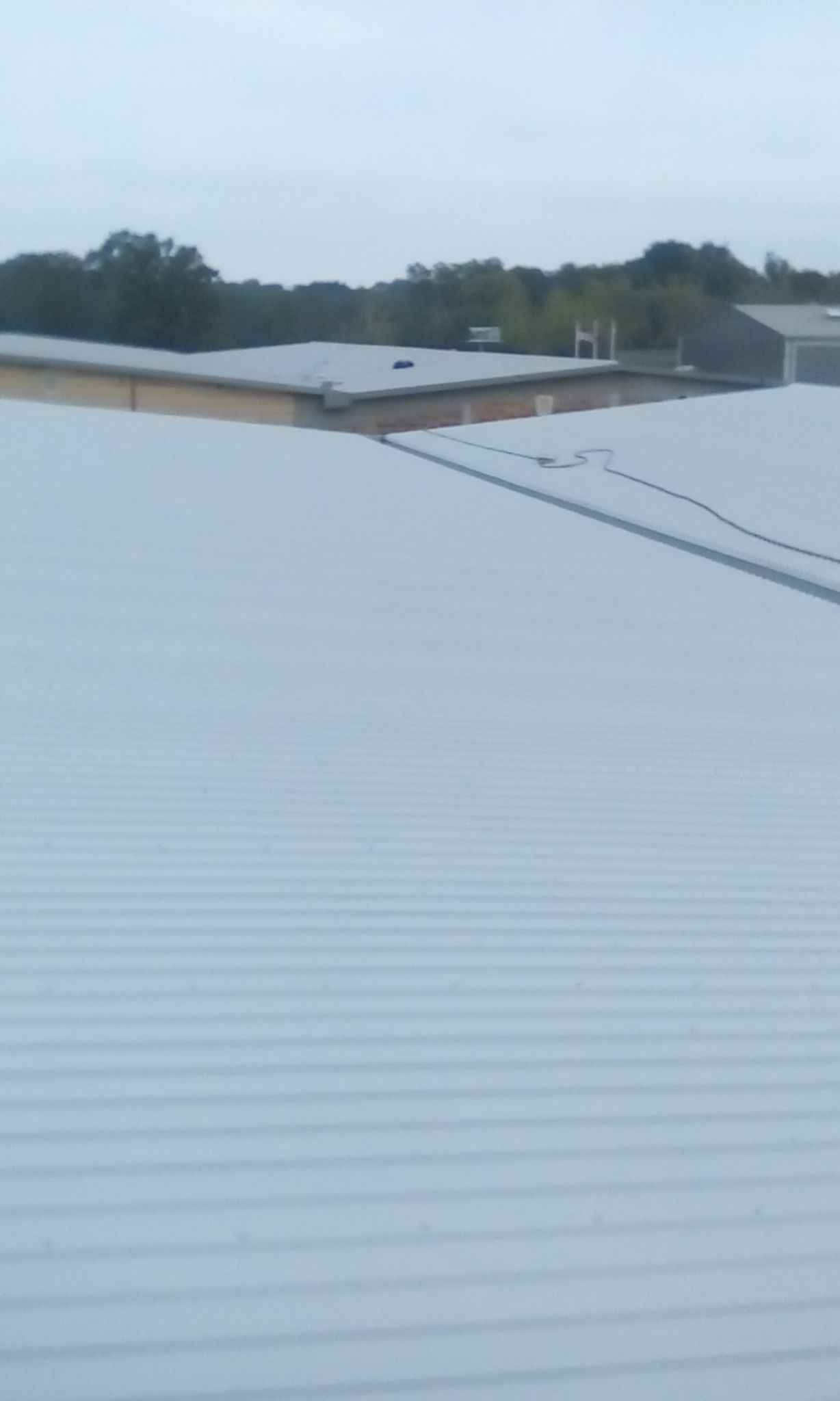 Widok z góry na nowo położone, jasnoszare, blaszane pokrycie dachowe z widocznymi liniami łączeń, na tle zabudowań i drzew w pochmurny dzień.