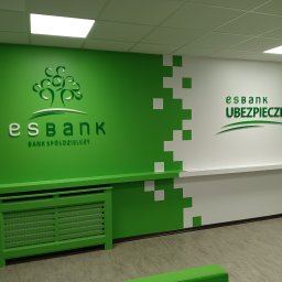 GRZEGORZ SOWIŃSKI FIRMA HANDLOWO USŁUGOWA INOWEST - Wnętrze biura z zielono-białą ścianą, logo 'esbank' i motywem drzewa, oraz napisem 'UBEZPIECZENIA' po prawej stronie. Grzejnik i listwa przypodłogowa również w kolorze zielonym.