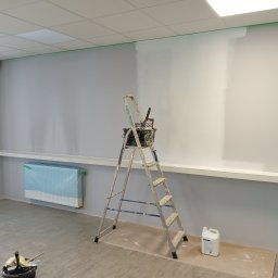 GRZEGORZ SOWIŃSKI FIRMA HANDLOWO USŁUGOWA INOWEST - Wnętrze biura w trakcie odświeżania: ściana pomalowana częściowo na biało, zabezpieczony grzejnik, drabina z narzędziami malarskimi i wiadro z farbą.