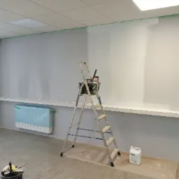 Wnętrze biura w trakcie odświeżania: ściana pomalowana częściowo na biało, zabezpieczony grzejnik, drabina z narzędziami malarskimi i wiadro z farbą.