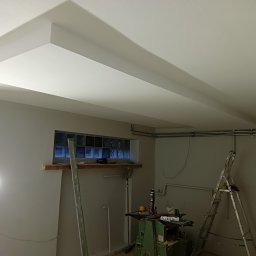 GRZEGORZ SOWIŃSKI FIRMA HANDLOWO USŁUGOWA INOWEST - Pomieszczenie w trakcie prac wykończeniowych, widoczny sufit z dekoracyjnym podwieszanym elementem, drabina oparta o ścianę, okno z luksferami oraz narzędzia i materiały budowlane.