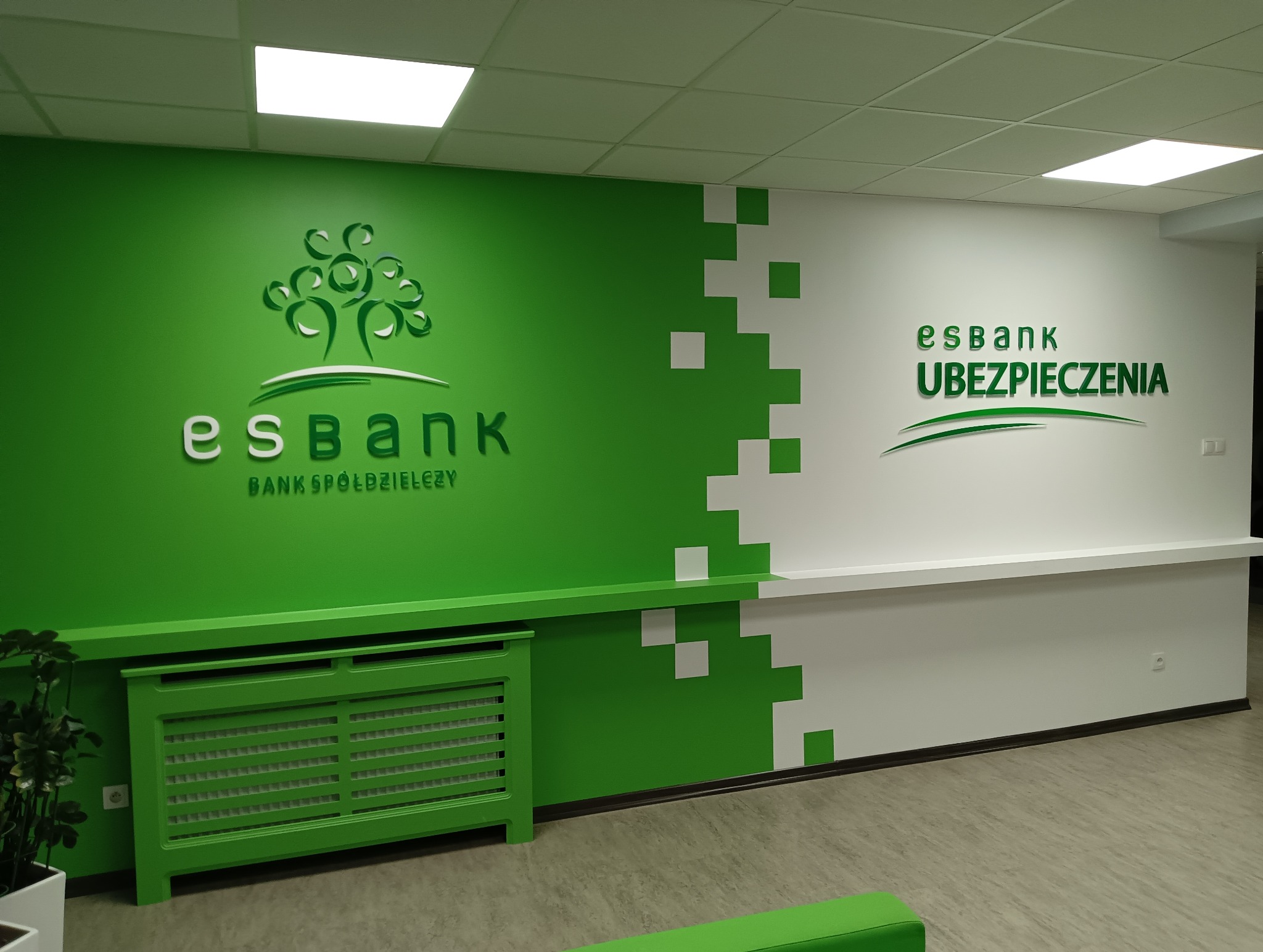 Wnętrze biura z zielono-białą ścianą, logo 'esbank' i motywem drzewa, oraz napisem 'UBEZPIECZENIA' po prawej stronie. Grzejnik i listwa przypodłogowa również w kolorze zielonym.