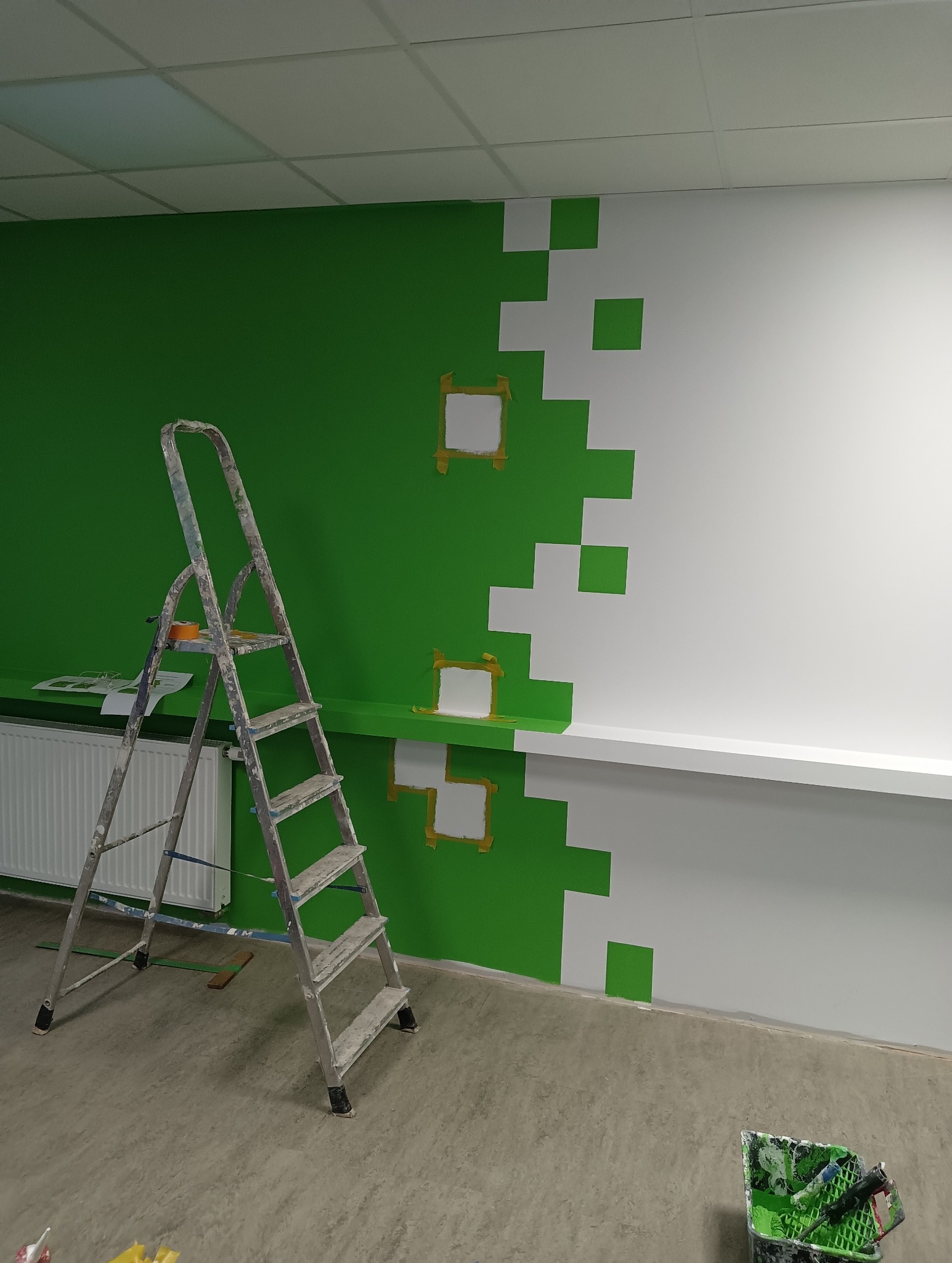 Ściana w biurze w trakcie malowania na zielono i biało w pikselowy wzór, zabezpieczona taśmą malarską, drabina malarska obok, widoczny grzejnik i wiadro z farbą na podłodze.