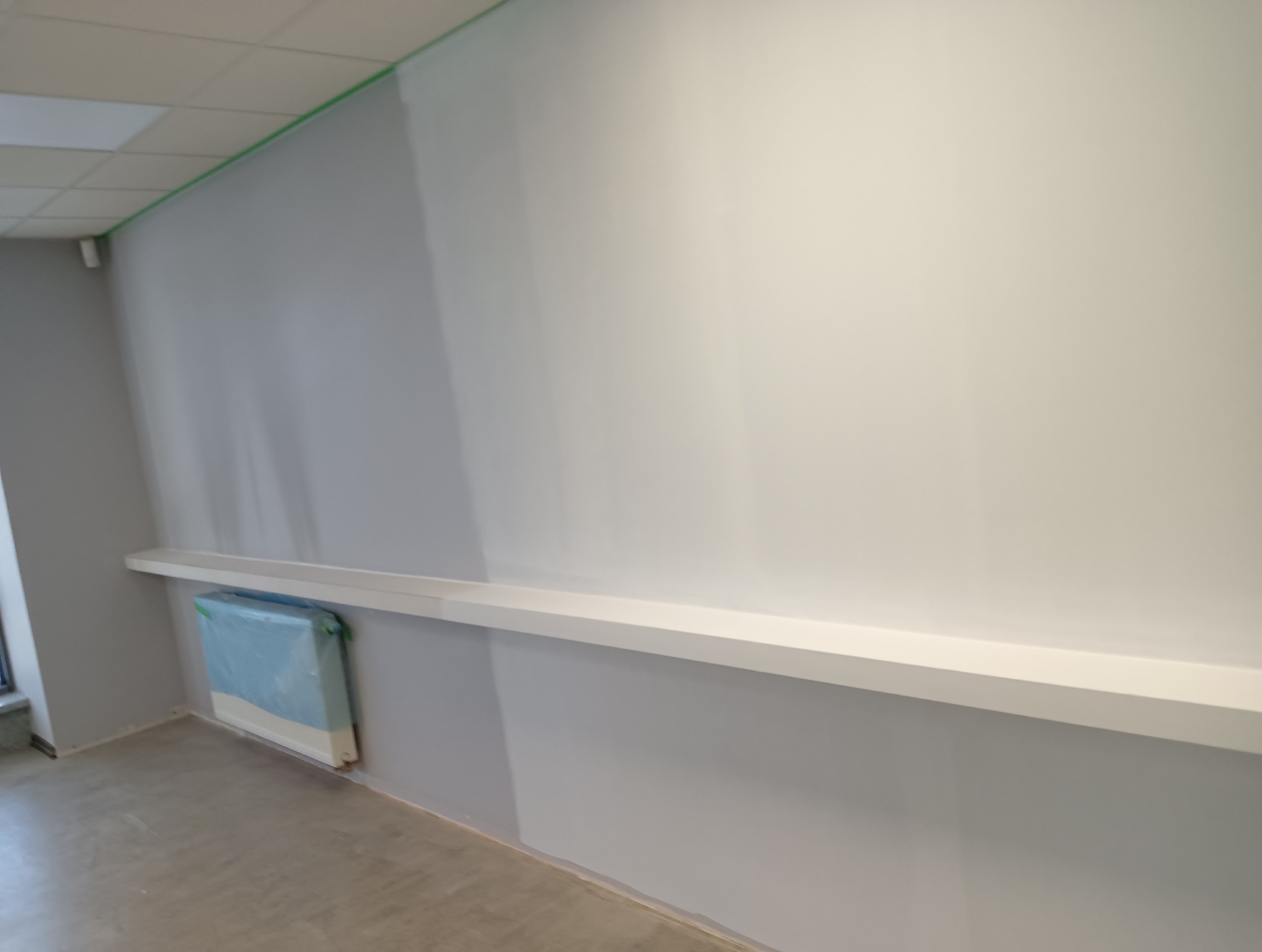 Świeżo pomalowane biuro z szarą ścianą w połowie wysokości, białą górną częścią i listwą przy suficie zabezpieczoną taśmą malarską, grzejnik zabezpieczony folią ochronną.