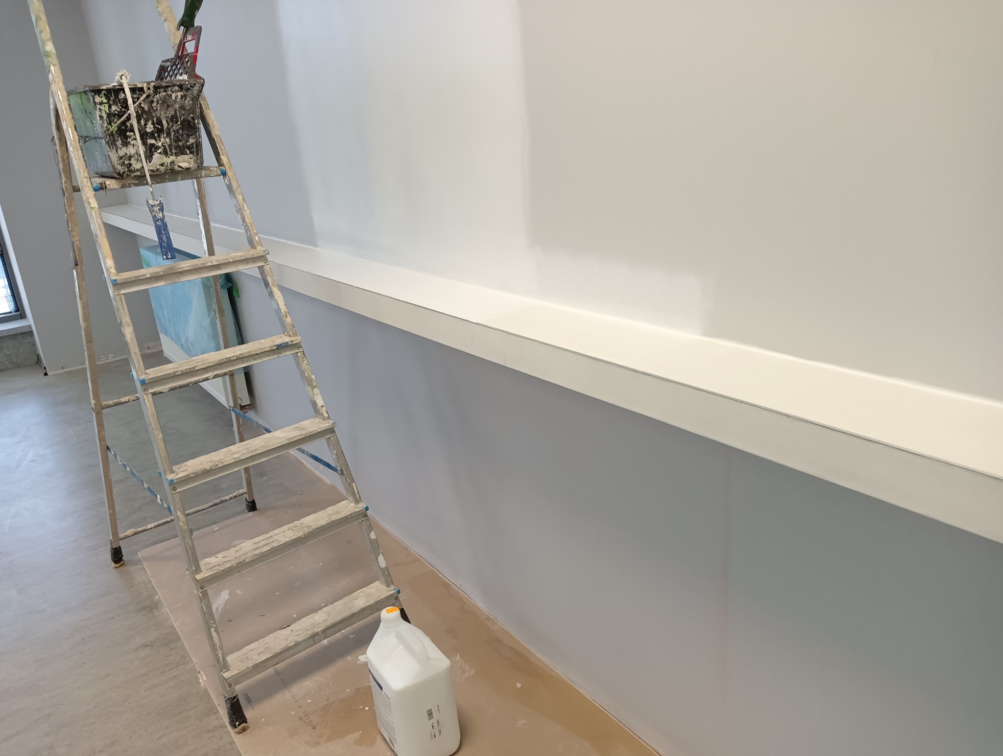 Świeżo pomalowana, jasnoszara ściana z białą listwą przypodłogową. Obok stoi aluminiowa drabina z wiadrem i narzędziami malarskimi, a na podłodze biały pojemnik po farbie.