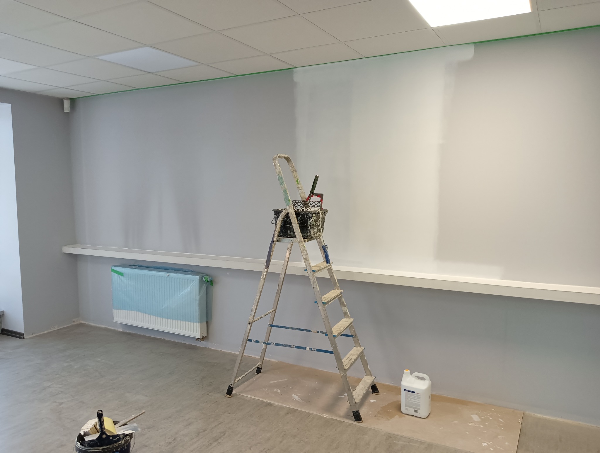 Wnętrze biura w trakcie odświeżania: ściana pomalowana częściowo na biało, zabezpieczony grzejnik, drabina z narzędziami malarskimi i wiadro z farbą.