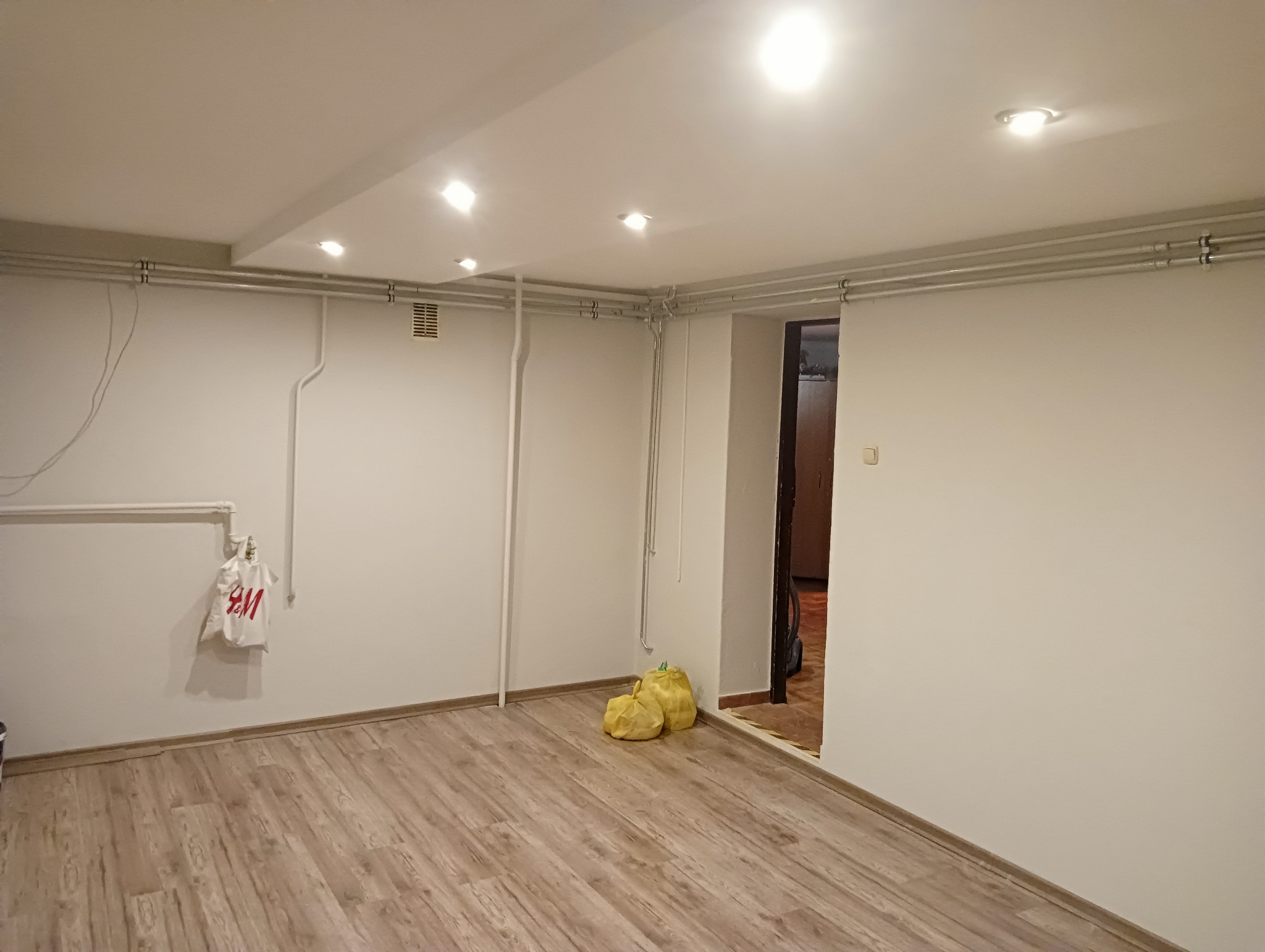 Puste, odświeżone wnętrze pokoju z panelami podłogowymi, świeżo pomalowanymi białymi ścianami i widocznymi rurami instalacyjnymi pod sufitem; na ścianie wisi torba z logo H&M, a w rogu leżą dwa...