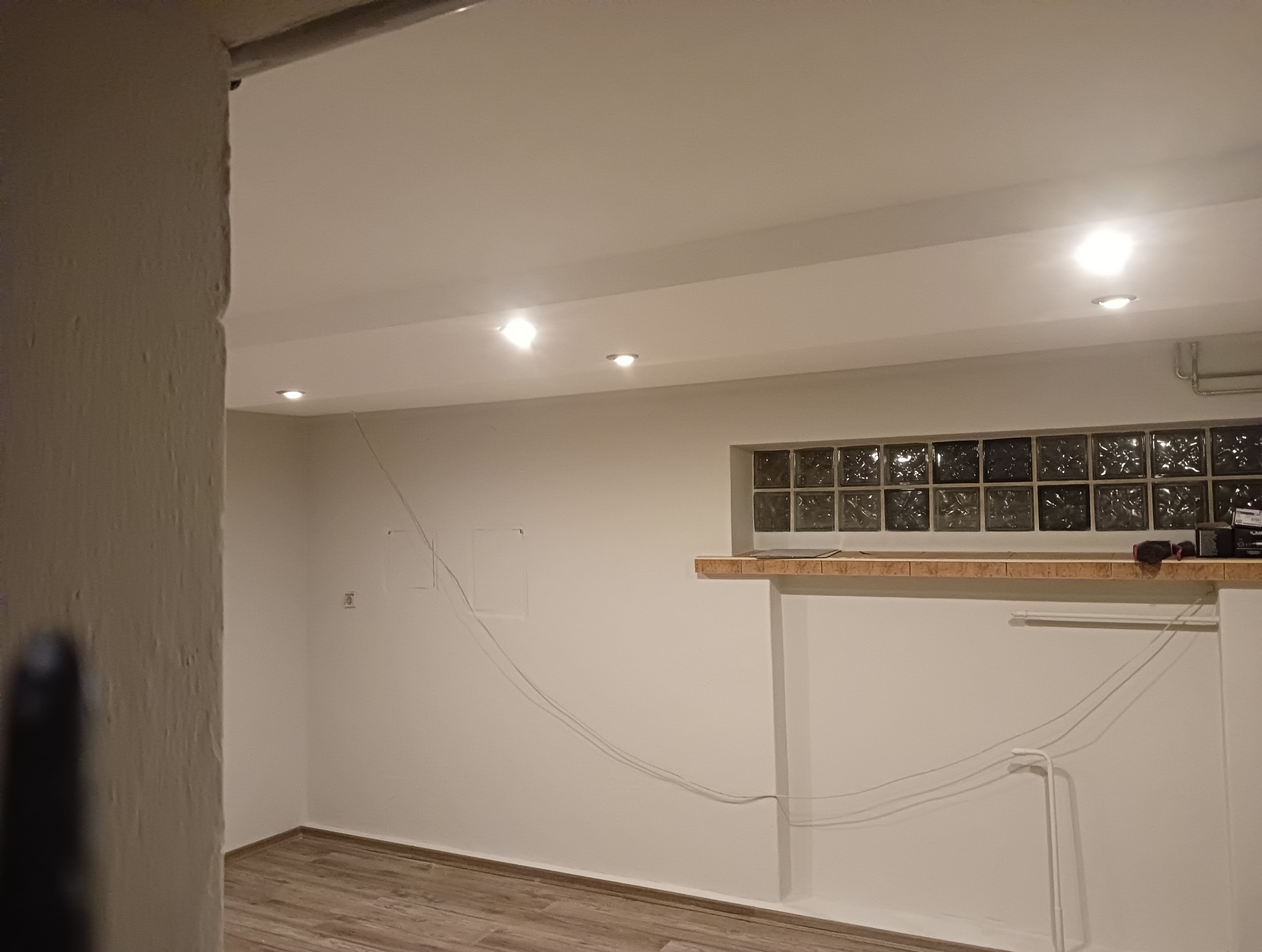 Surowe wnętrze piwnicy z oknem z luksferów, widoczne instalacje elektryczne i rury, podłoga wyłożona panelami imitującymi drewno.