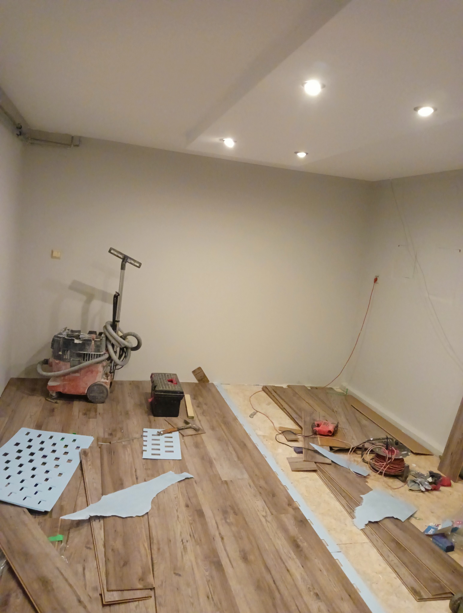 Układanie paneli podłogowych w pomieszczeniu: widoczny częściowo ułożony parkiet, narzędzia, odkurzacz przemysłowy i resztki materiałów wykończeniowych.