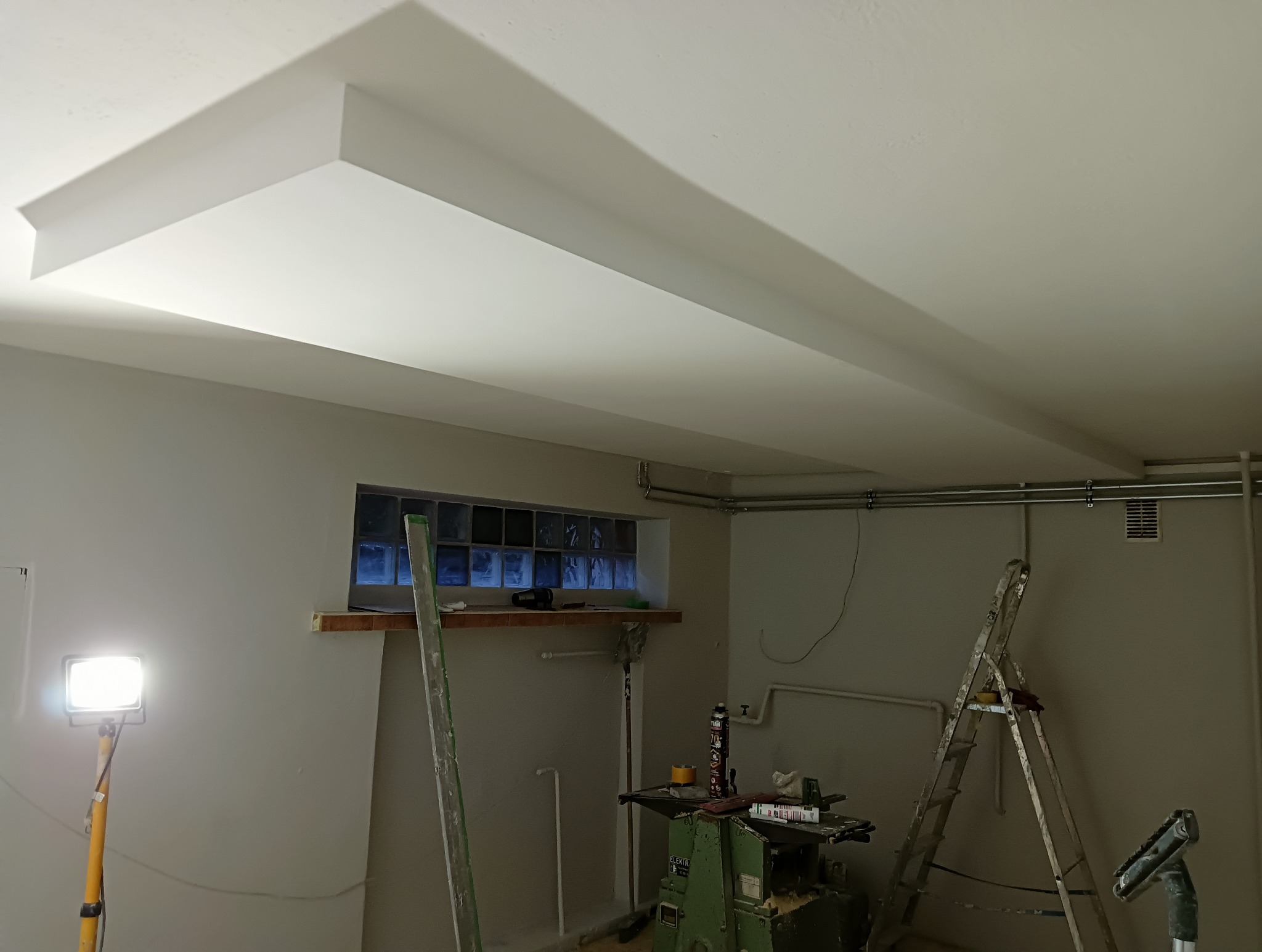 Pomieszczenie w trakcie prac wykończeniowych, widoczny sufit z dekoracyjnym podwieszanym elementem, drabina oparta o ścianę, okno z luksferami oraz narzędzia i materiały budowlane.