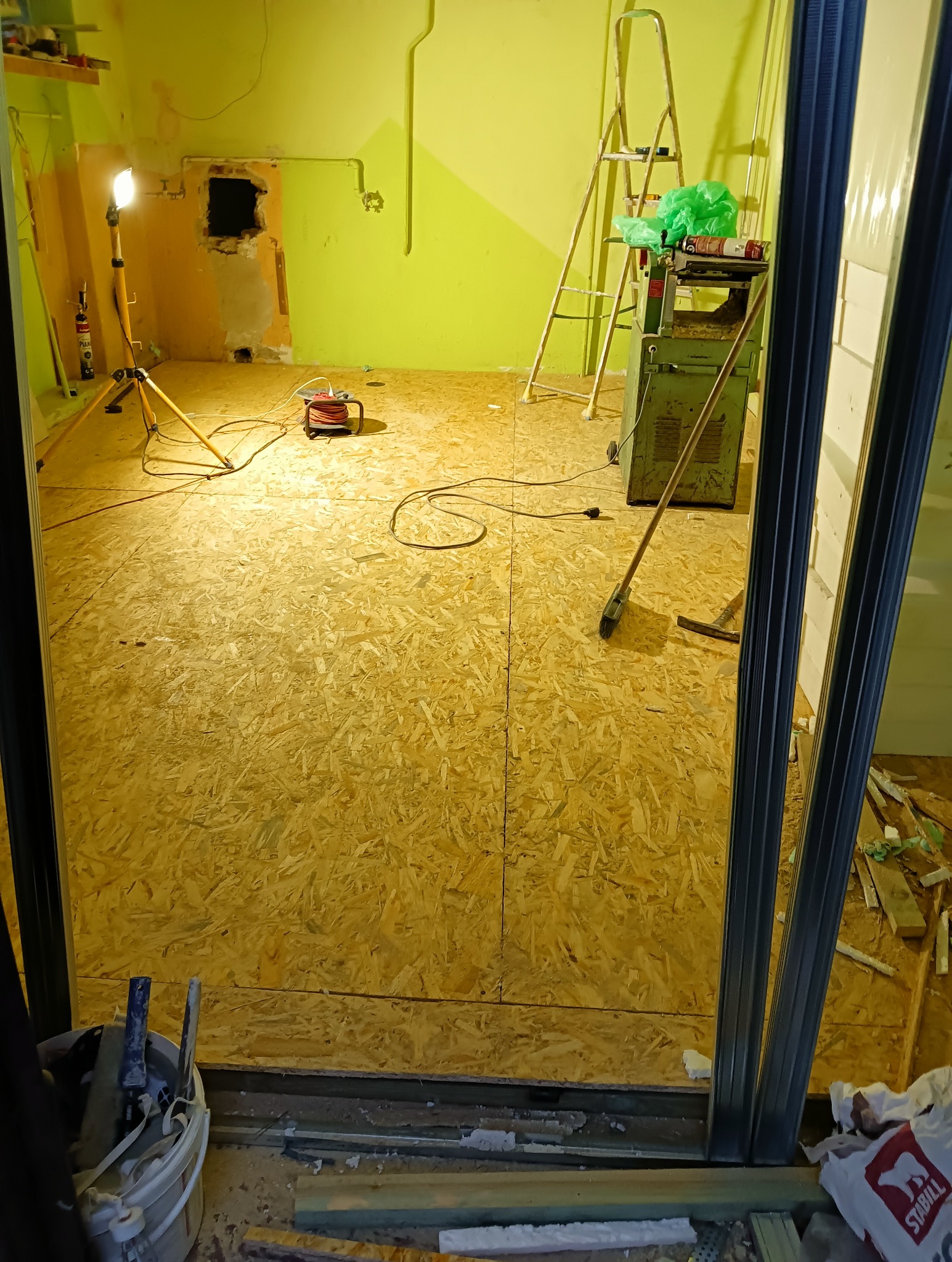 Wnętrze w trakcie remontu z nową podłogą z płyt OSB, widoczne narzędzia, drabina, lampa robocza i maszyna do obróbki drewna na tle limonkowych ścian.