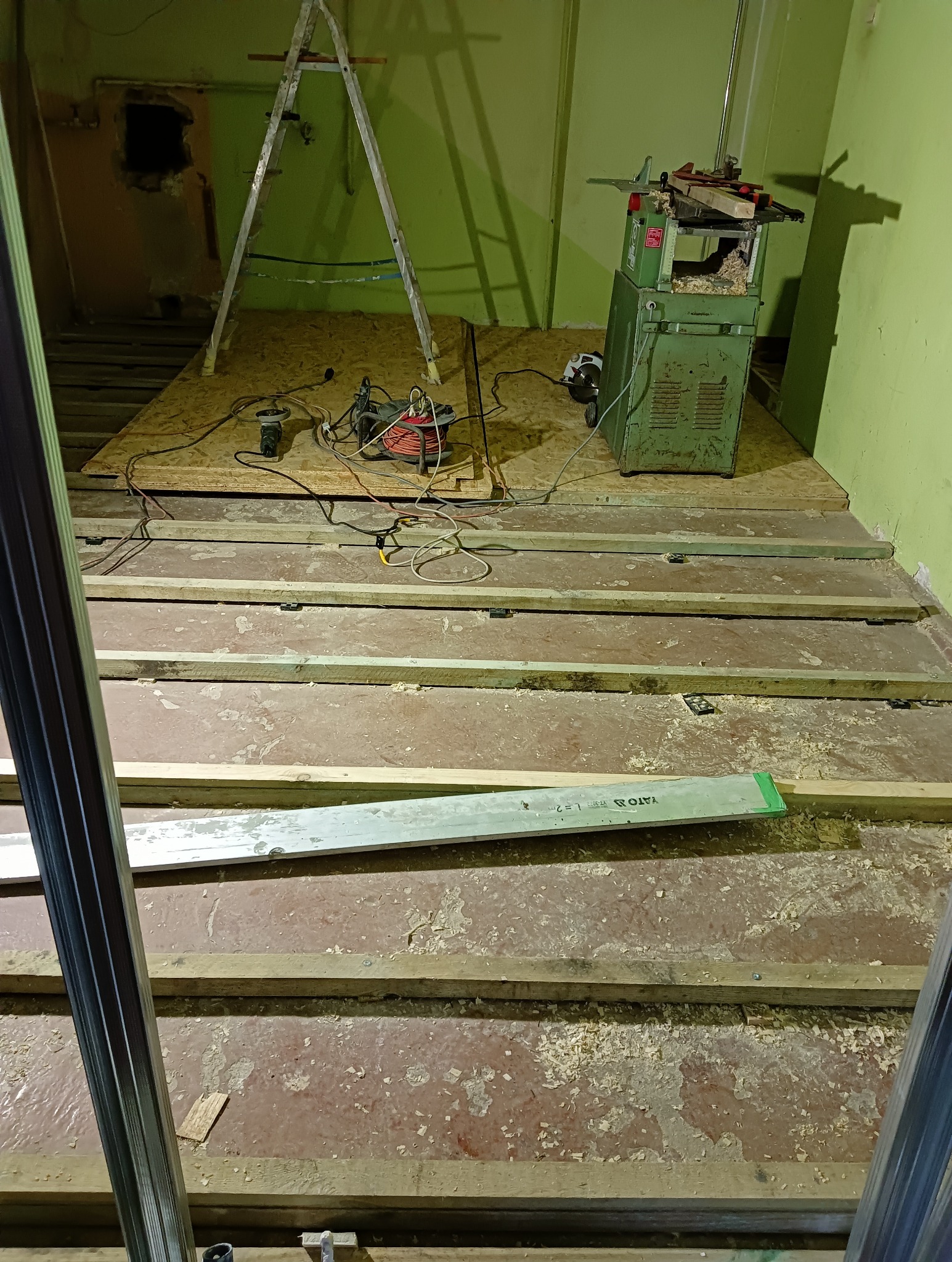 Przygotowanie do ułożenia podłogi: widoczne legary na starym podłożu, narzędzia, poziomica YATO i strugarka na tle zielonej ściany.