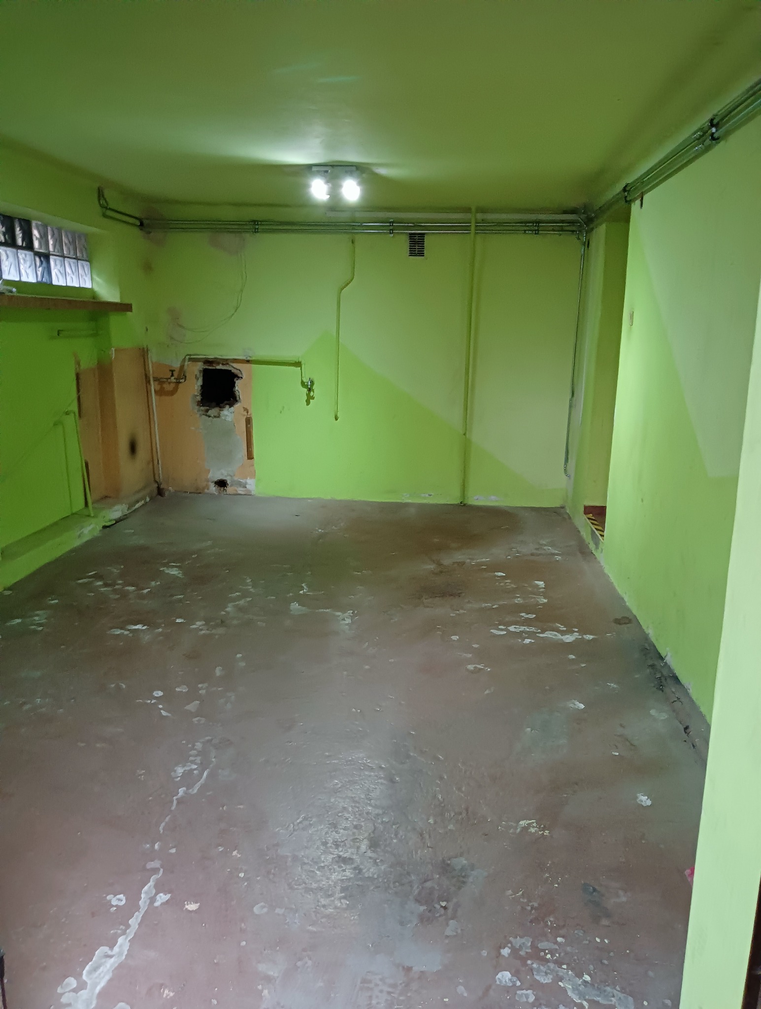 Pusty pokój w odcieniach zieleni z uszkodzeniami ściany, widoczne rury pod sufitem i ślady po zalaniu na podłodze.