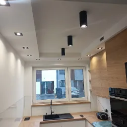 Nowoczesna kuchnia z białymi szafkami, drewnianym blatem i czarnym zlewem pod oknem. Na suficie zamontowane punktowe i tubowe oświetlenie.