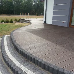 Brązowy taras z desek kompozytowych, wykończony obrzeżem z szarej kostki brukowej, prowadzący do schodów z kostki i budynku z szarą elewacją, widok z boku.