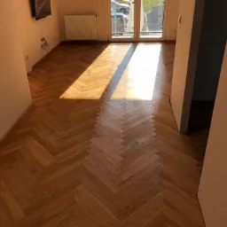 Ułożony parkiet w jodełkę w jasnym, pustym pokoju z padającym promieniem słońca, widoczne okno balkonowe i grzejnik.