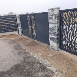 Nowoczesna, grafitowa brama wjazdowa z ozdobnymi wycięciami, zamontowana na słupkach z szarej kostki, uzupełniona o furtkę z identycznym wzorem, widoczna na tle szarego nieba w Grucznie.