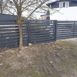 Nowoczesne, poziome ogrodzenie panelowe w kolorze grafitowym z bramą wjazdową, widoczne z zewnątrz posesji, z drzewem bez liści po lewej stronie i białym domem w tle.