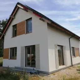 Dwukondygnacyjny dom jednorodzinny z białą elewacją, drewnianymi panelami dekoracyjnymi i żaluzjami okiennymi, w tle las i błękitne niebo, widoczne profile metalowe przy ziemi.