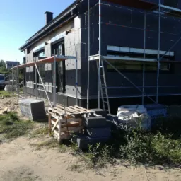 Dom w trakcie ocieplania, widoczne rusztowania, czarny styropian elewacyjny, paleta i paczki styropianu na ziemi.