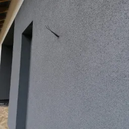 Szara elewacja domu z widocznym fragmentem okna i wystającymi kablami, wykończona tynkiem strukturalnym typu baranek.