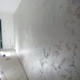 Ściana z dekoracyjną, nieregularną fakturą w odcieniach szarości i beżu, widoczna podczas procesu wykańczania, z widocznymi przewodami i narzędziami na podłodze.