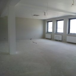 Firma remontowa - Wnętrze pustego, jasnego pomieszczenia w trakcie prac wykończeniowych. Widoczne białe ściany, surowy beton na podłodze, grzejniki pod oknami i wystające przewody z sufitu. Charakterystyczny, gruby...