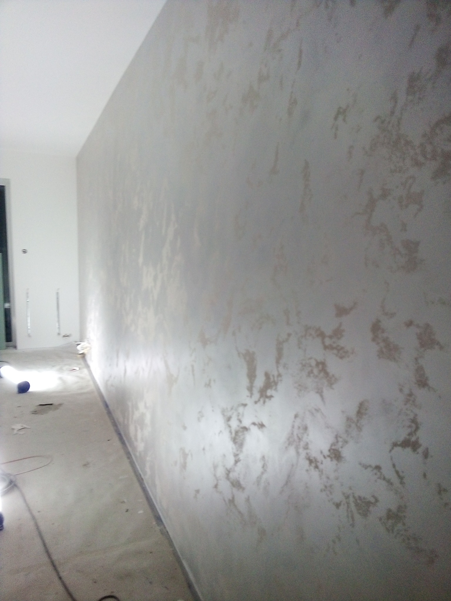 Ściana z dekoracyjną, nieregularną fakturą w odcieniach szarości i beżu, widoczna podczas procesu wykańczania, z widocznymi przewodami i narzędziami na podłodze.