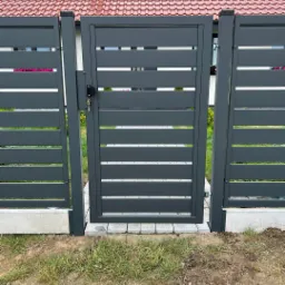 Nowoczesna, grafitowa brama wjazdowa z poziomymi panelami, zamontowana między dwoma segmentami ogrodzenia panelowego na betonowej podmurówce i kostce brukowej, widok z zewnątrz posesji.