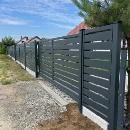 Nowoczesne, poziome przęsła w antracytowej kolorystyce, tworzące ogrodzenie posesji z przesuwną bramą, widoczne na tle błękitnego nieba i zieleni ogrodu.