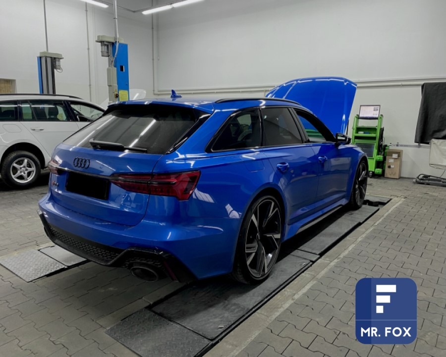 Niebieskie Audi RS6 Avant z otwartą maską w warsztacie, widoczny podnośnik i sprzęt diagnostyczny, logo firmy w prawym dolnym rogu.