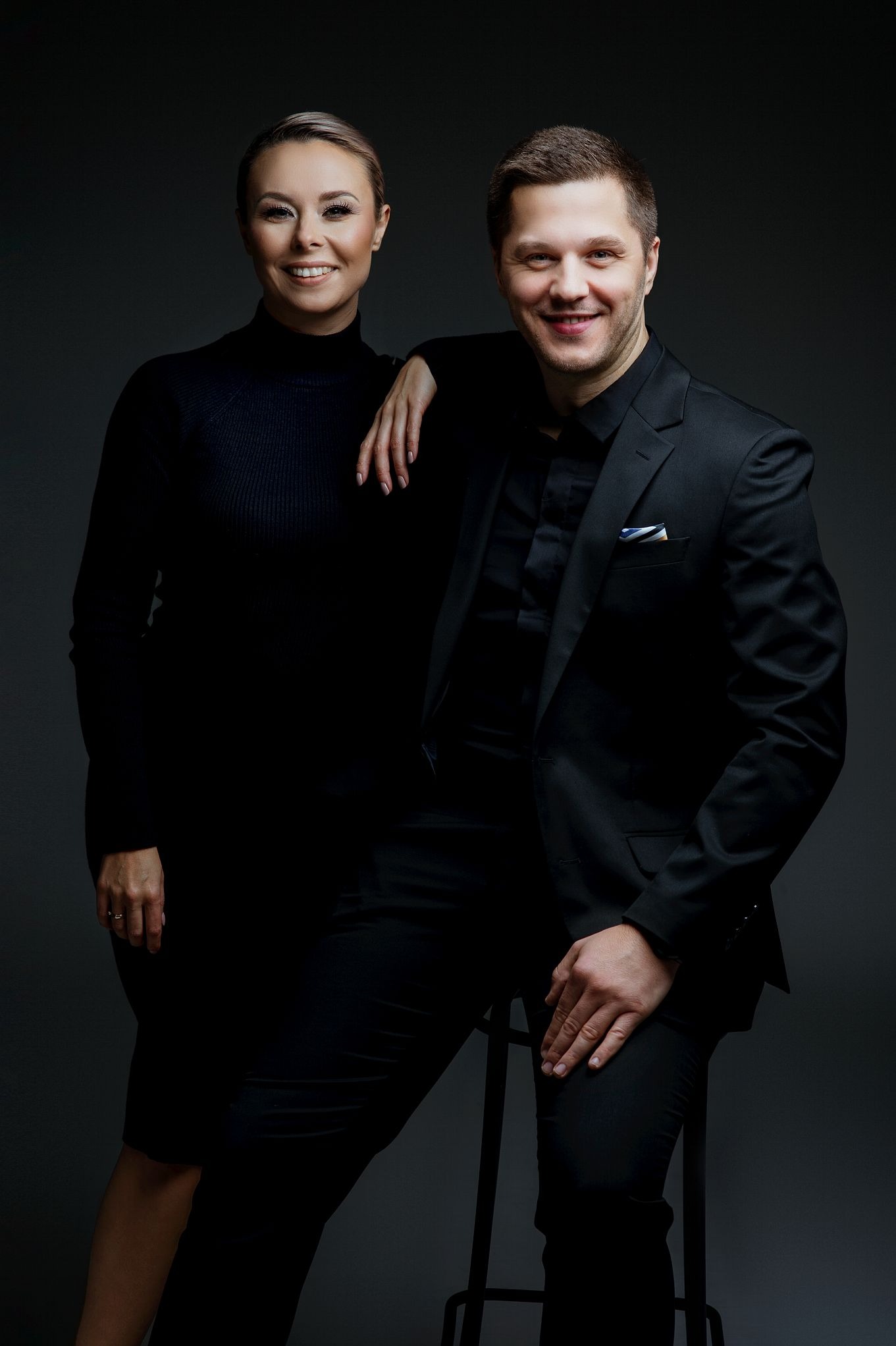 Portret kobiety opierającej rękę na ramieniu mężczyzny siedzącego na stołku, oboje ubrani na czarno, z uśmiechem na twarzy, na ciemnoszarym tle.