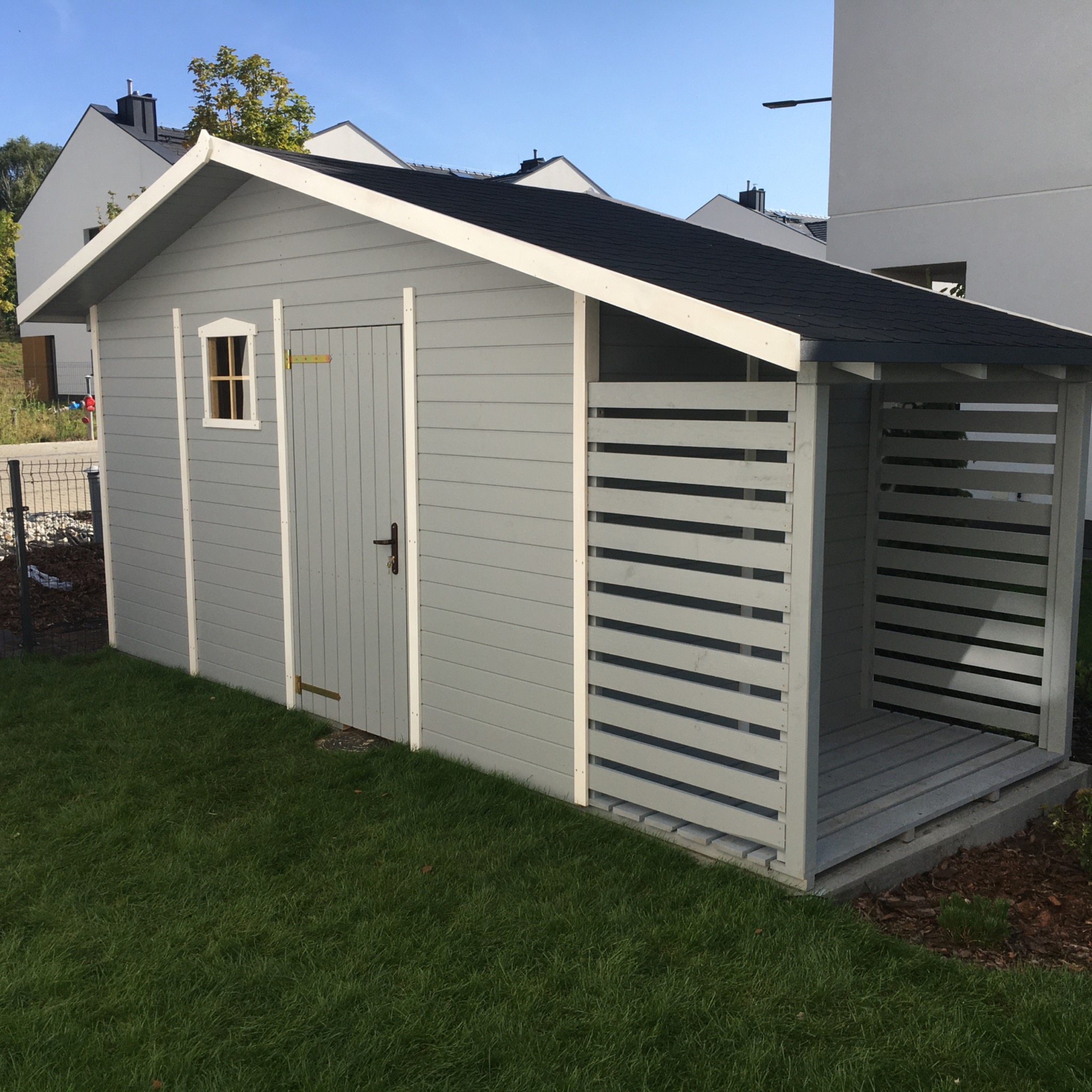 Szara drewniana drewutnia z zamykanymi drzwiami i otwartą częścią na drewno, na tle zielonego trawnika i nowoczesnych domów.