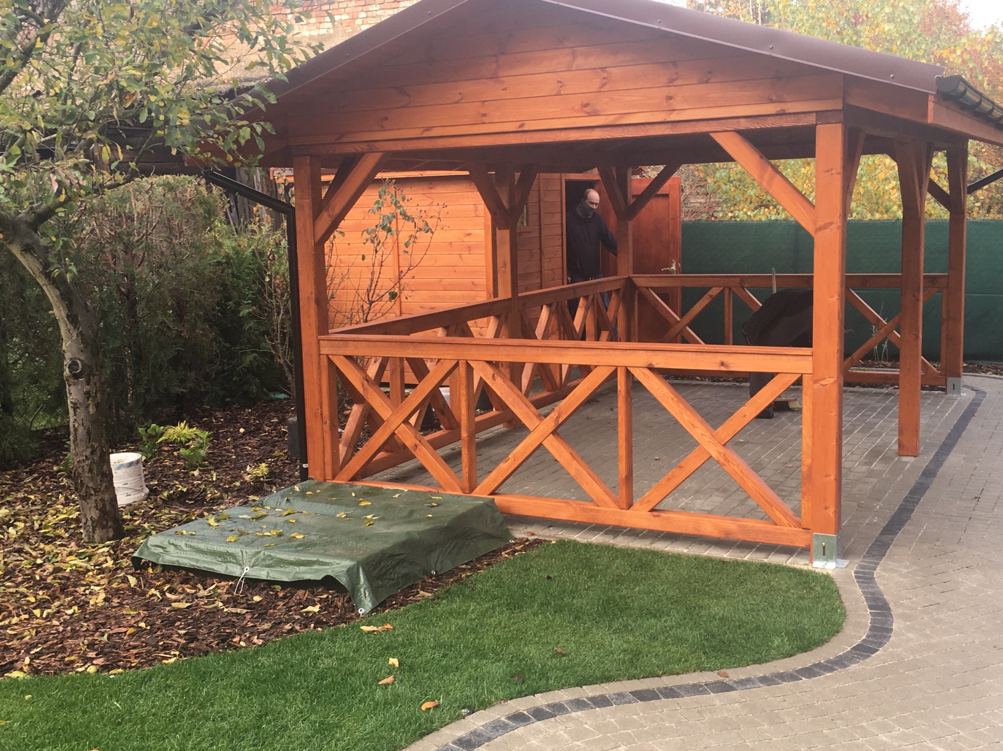 Drewniana altana z brązowym dachem na posesji, widoczne elementy konstrukcyjne i wykończeniowe, w tle budynek gospodarczy z otwartymi drzwiami i osoba w środku.