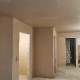 Wnętrze w trakcie prac wykończeniowych: ściany i sufit pokryte świeżo nałożoną, jasną gładzią, widoczne surowe ościeżnice drzwiowe i narzędzia budowlane w tle.