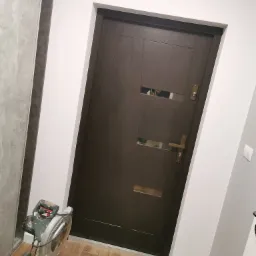 Ciemne drzwi wewnętrzne z dwoma poziomymi przeszkleniami, zamontowane w białej futrynie, na podłodze panele, w tle widoczna piła ukośnica.