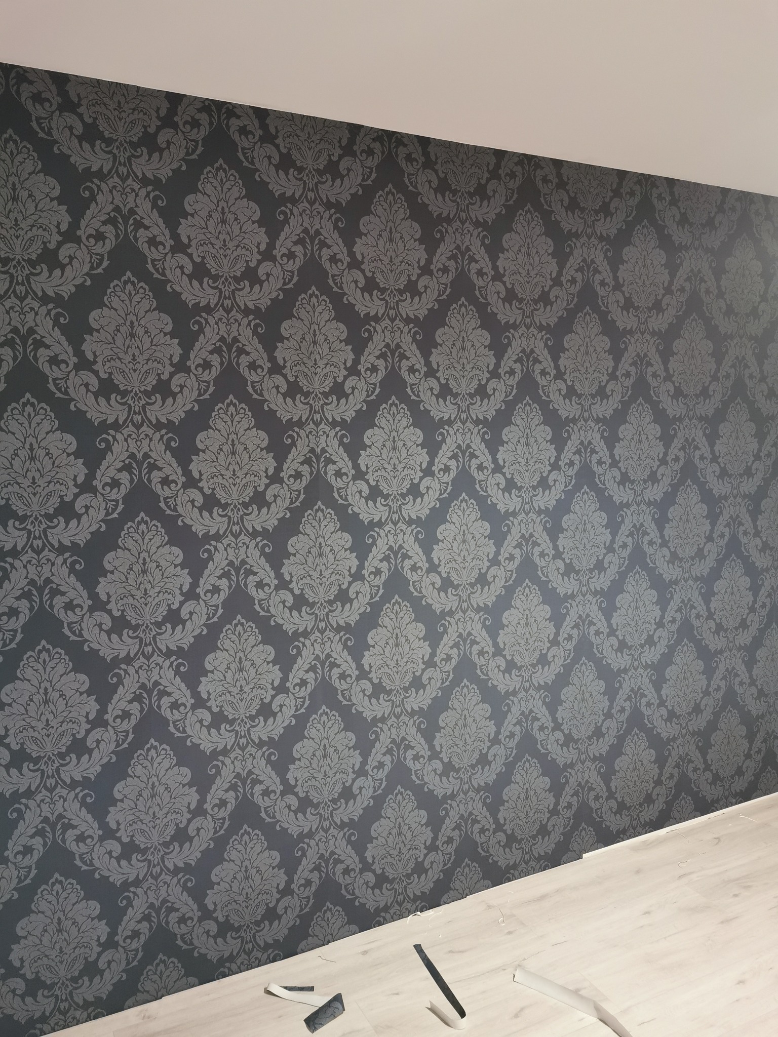 Ściana pokryta ciemnoszarą tapetą w klasyczny, roślinny wzór; fragmenty tapety i podłogi widoczne w dolnym rogu.