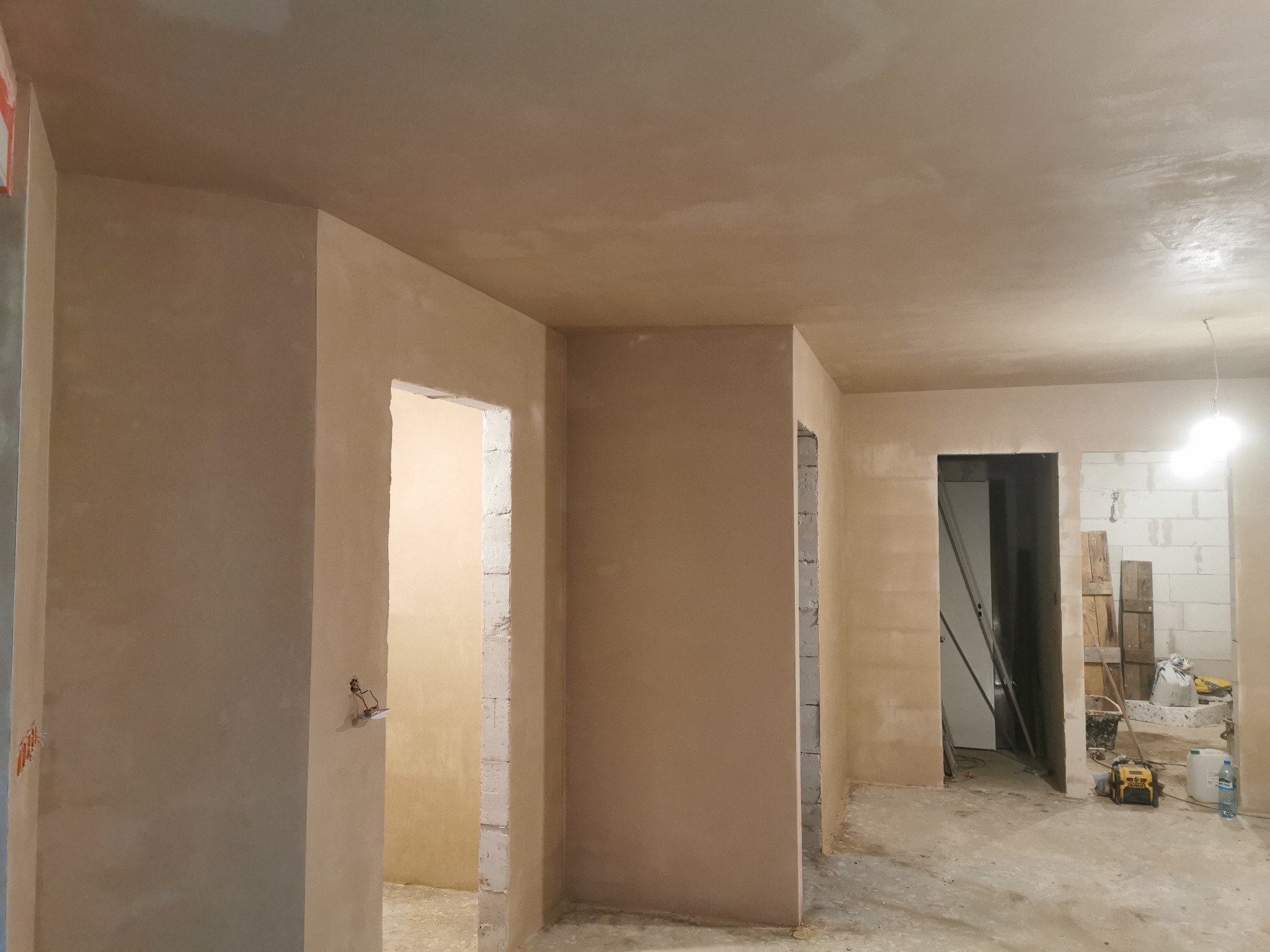Wnętrze w trakcie prac wykończeniowych: ściany i sufit pokryte świeżo nałożoną, jasną gładzią, widoczne surowe ościeżnice drzwiowe i narzędzia budowlane w tle.