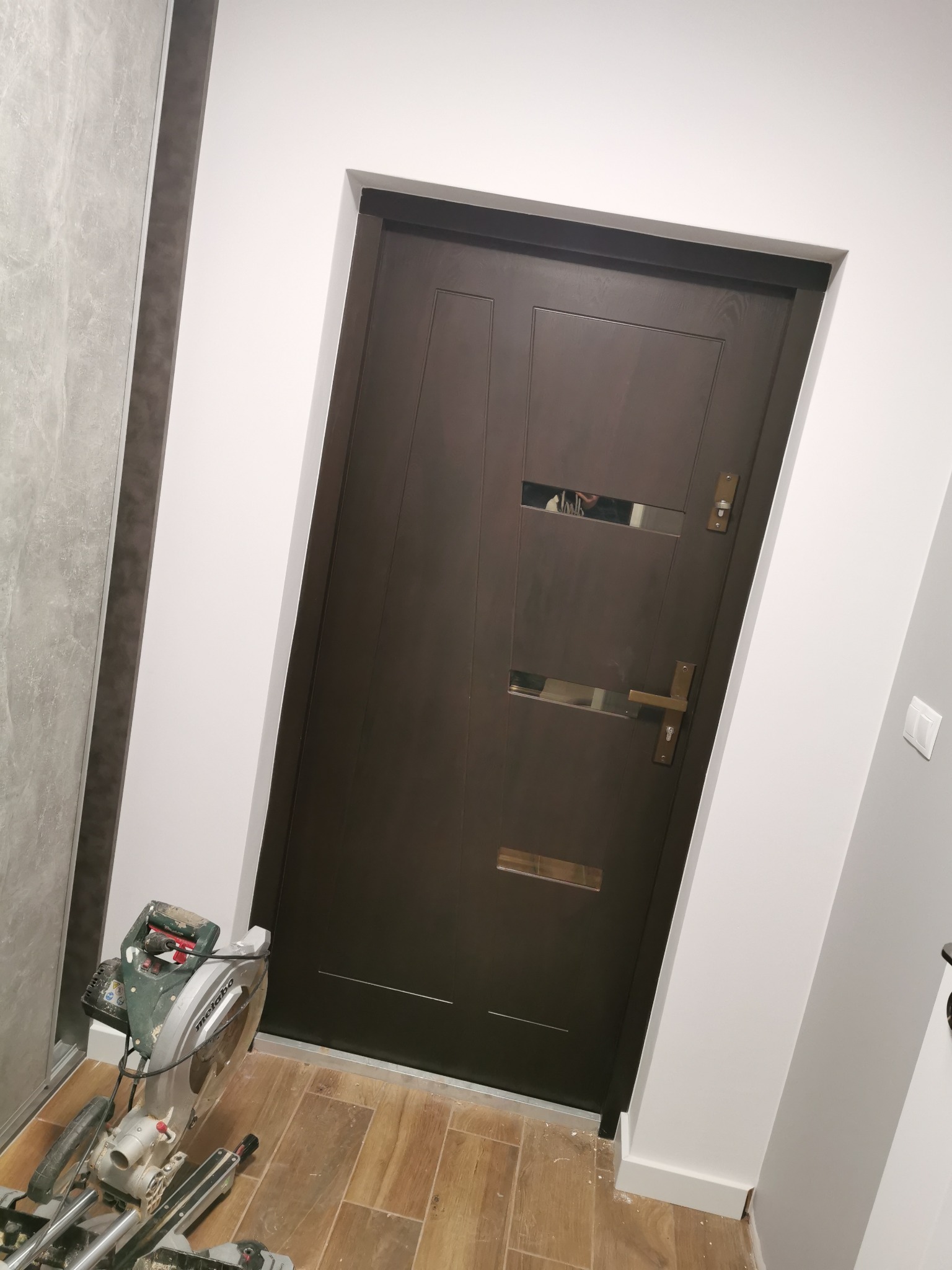 Ciemne drzwi wewnętrzne z dwoma poziomymi przeszkleniami, zamontowane w białej futrynie, na podłodze panele, w tle widoczna piła ukośnica.