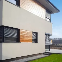 Nowoczesny dom z jasną elewacją i drewnianymi panelami, okna zabezpieczone szarymi systemami osłonowymi, częściowo opuszczonymi, widoczne logo producenta w rogu zdjęcia.