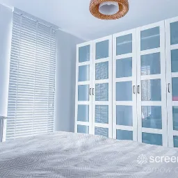 Jasne, poziome żaluzje z drewna w oknie sypialni, widoczne na tle białej szafy z przeszklonymi drzwiami i wzorzystej pościeli.