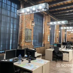 Biuro w stylu loft z dużymi oknami wyposażonymi w pionowe żaluzje w kolorze antracytowym, oświetlenie sufitowe w metalowych obudowach, ściany z cegły.