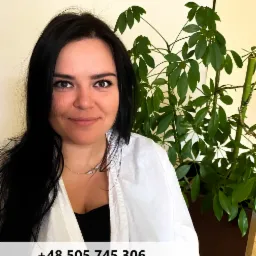 Portret uśmiechniętej kobiety z ciemnymi włosami, w białym ubraniu, na tle rośliny doniczkowej.