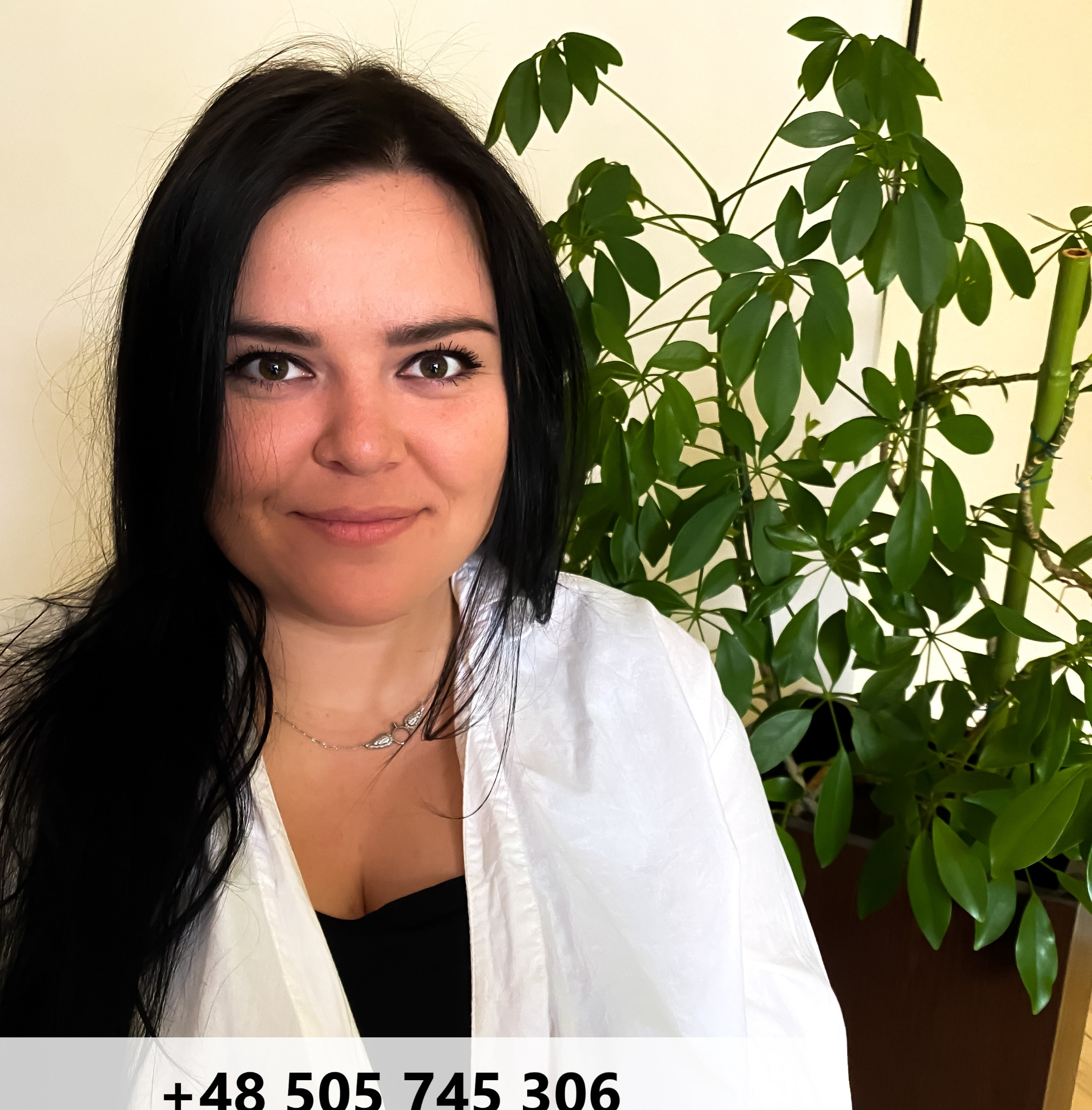 Portret uśmiechniętej kobiety z ciemnymi włosami, w białym ubraniu, na tle rośliny doniczkowej.