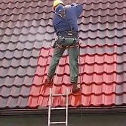 Pracownik w kasku i szelkach bezpieczeństwa czyści dachówkę z drabiny, widoczna różnica w kolorze między wyczyszczoną a brudną częścią dachu.