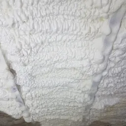 Świeżo nałożona, gęsta warstwa białej pianki izolacyjnej z widoczną strukturą komórkową, szczelnie wypełniająca przestrzeń poddasza, z drobnymi okruchami na drewnianej podłodze.